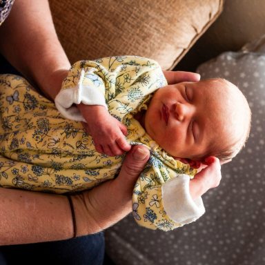 A person gently holds a sleeping newborn in a patterned yellow outfit.