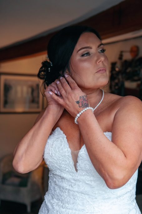 A bride in a white gown adjusting her earrings, showcasing a tattoo on her hand.