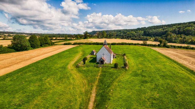A small white chapel surrounded by green fields and scattered trees under a blue sky.