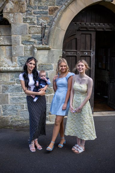 Three young women stand together, one holding a baby, in front of a church doorway.
