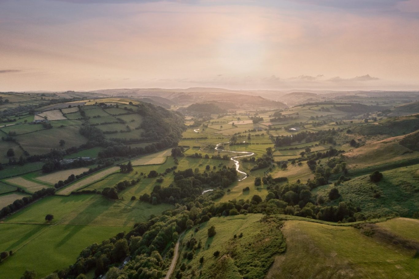 A scenic landscape featuring rolling hills, fields, and a winding river under a soft sunset glow.