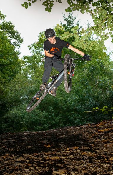 A cyclist performing a trick in mid-air above a dirt path surrounded by trees.