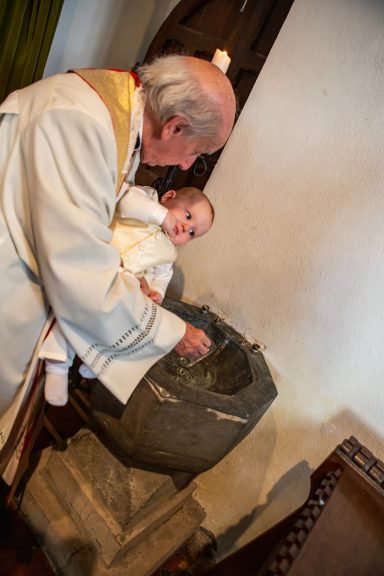 A priest holds a baby over a stone font in a church for a baptism ceremony.