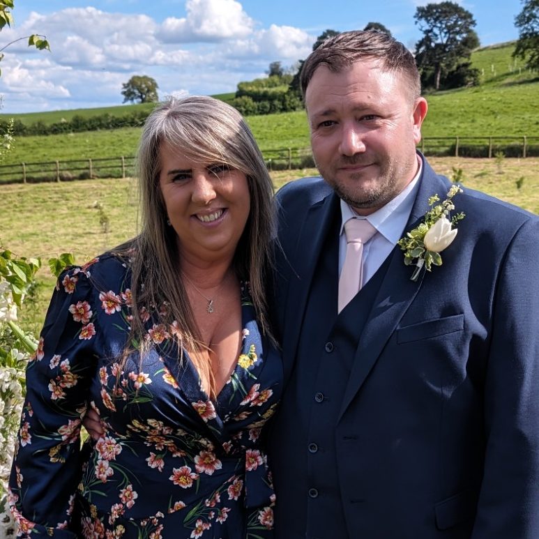 A couple poses outdoors, surrounded by greenery, smiling at the camera.