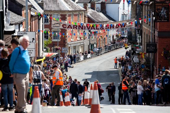 Crowds line a street adorned with bunting for a festival, with steep buildings on either side.