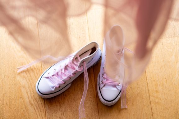 White Converse shoes with pink laces, set on a wooden floor, next to flowing fabric.