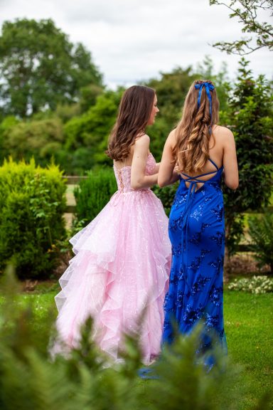 Two young women in elegant dresses, one pink and one blue, walking in a garden setting.