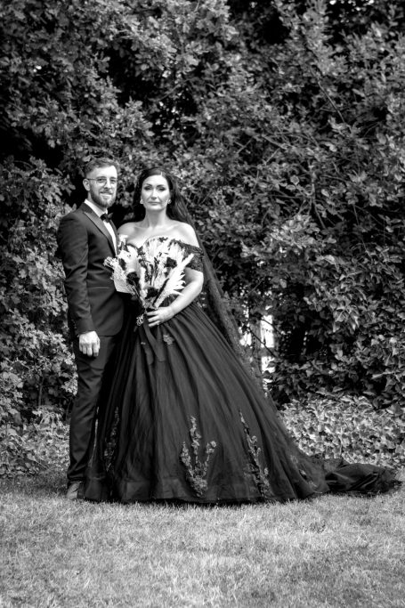 A couple in formal attire poses outdoors, surrounded by greenery.