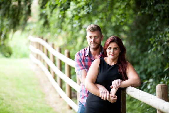 A couple stands smiling by a wooden fence in a green outdoor setting.