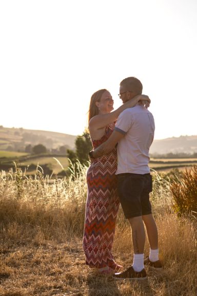 A couple embracing in a sunlit field, surrounded by greenery and hills.