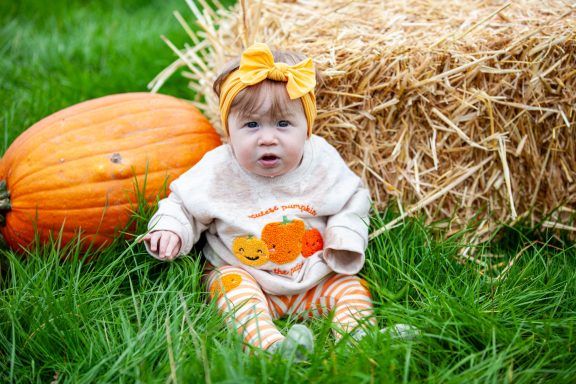 Baby sitting on grass beside a pumpkin and a hay bale, wearing a pumpkin-themed outfit.