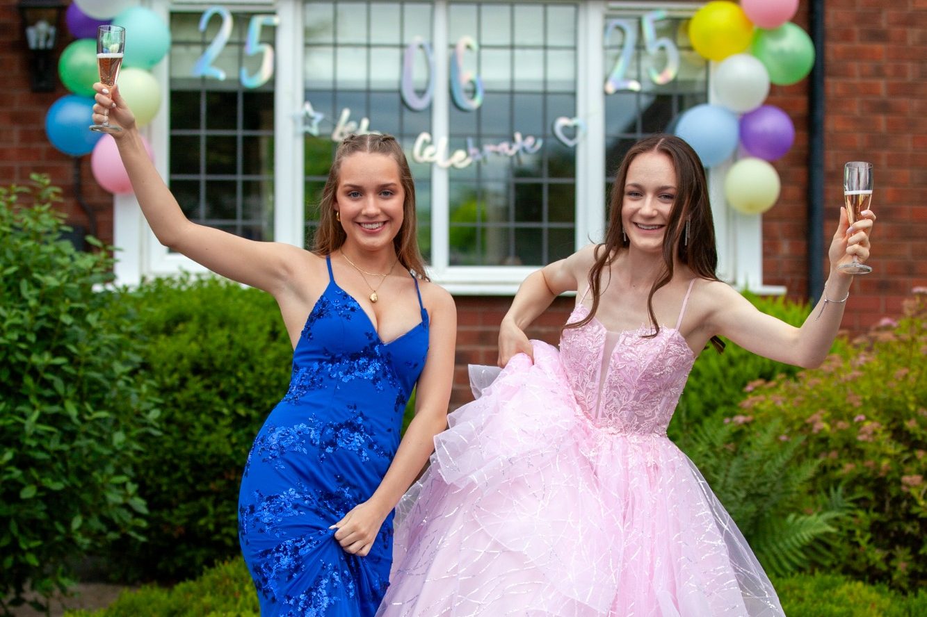 Two young women celebrate, one in a blue dress and the other in a pink gown, posing joyfully.