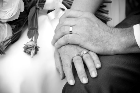 Two hands with wedding rings clasped together, resting on a white surface with a rose.