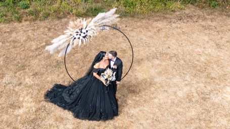 A couple in formal attire embraces under a decorative circular structure in a field.