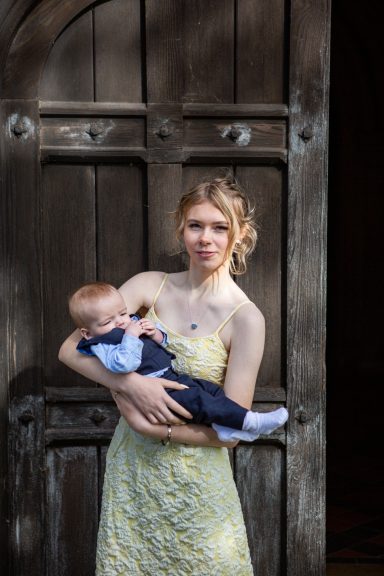 A young lady in a yellow dress holds a baby while standing by a wooden door.
