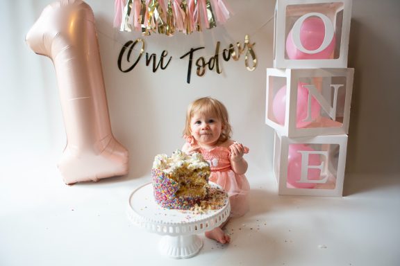 A baby girl in a pink dress smiles beside a birthday cake and decorative blocks spelling 'ONE'.