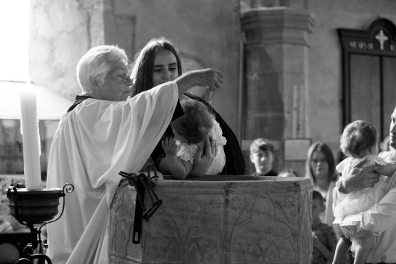 A minister baptises a child in a church, surrounded by family members.