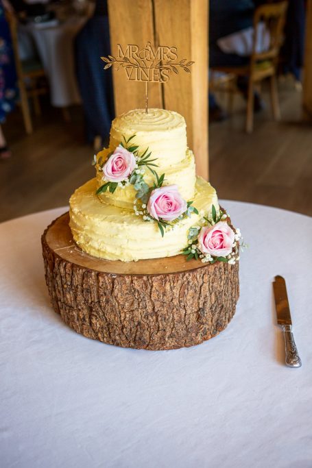 A three-tiered yellow cake adorned with pink roses, placed on a wooden base.