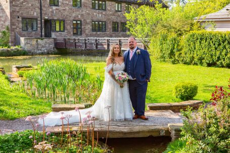 A bride and groom stand on a bridge over a pond in a lush garden setting.
