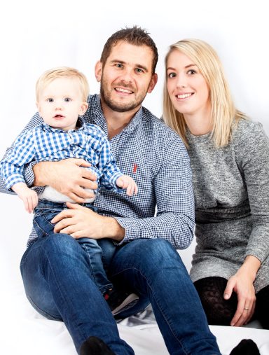 A family portrait featuring a couple with their baby, smiling against a white background.