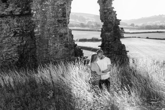 A couple stands among overgrown grass near crumbling stone ruins.