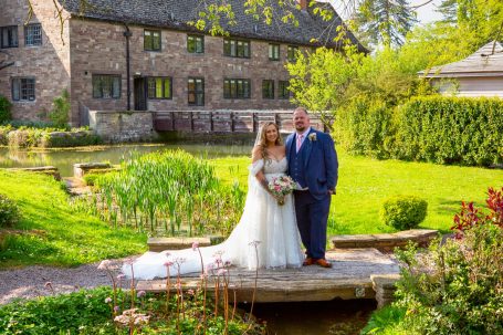 Bride in a wedding dress and groom in a suit standing on a bridge in a lush garden.