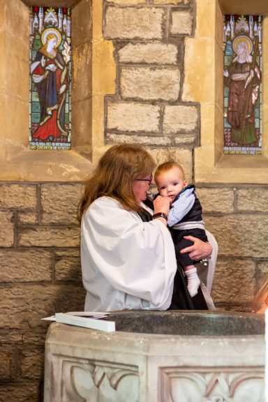A woman in a white robe holds a baby near a baptismal font in a church.