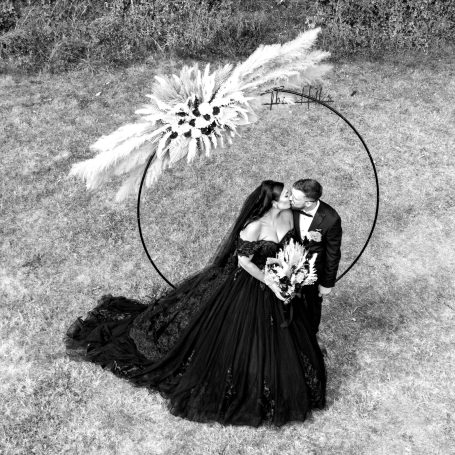 A couple in formal attire embraces, framed by a large floral hoop in a grassy setting.