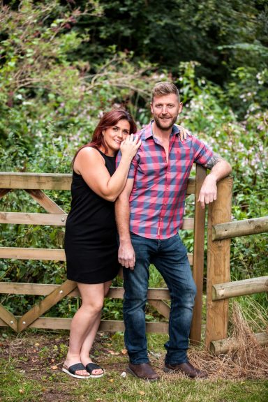 A couple stands by a wooden gate in a lush outdoor setting, smiling at the camera.