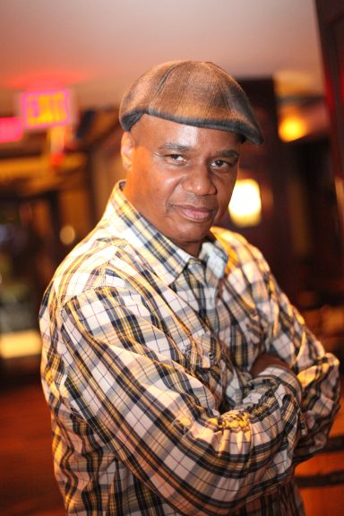 A man in a checkered shirt and cap poses confidently with arms crossed.