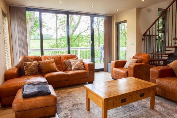 Modern living room with brown leather furniture and large windows overlooking greenery.