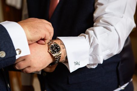 Two hands one helping to fasten a Rolex watch, both dressed in formal attire.