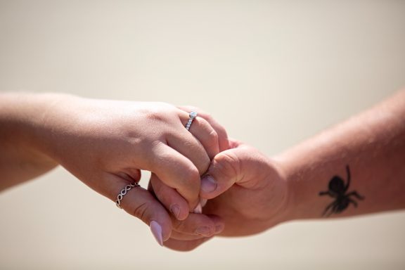 Two hands, one with rings, clasped together, with an ant tattoo visible on the wrist.