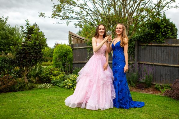 Two women in elegant evening gowns pose together in a garden setting.