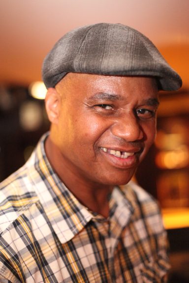 Smiling man wearing a grey flat cap and a checked shirt, with a warm background.