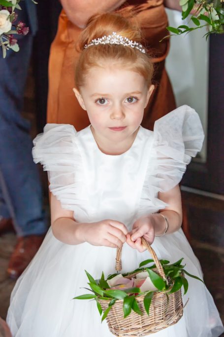Young girl in a white dress with a floral basket, sitting with a serious expression.