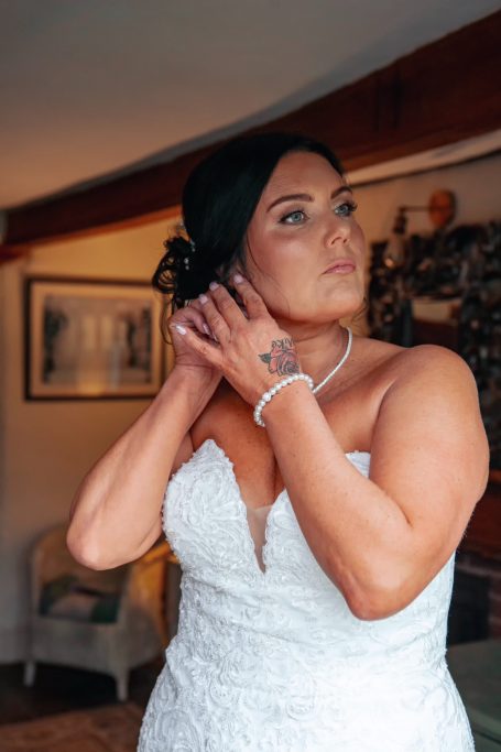 Bride adjusting her earrings in a softly lit room, wearing a lace wedding dress.