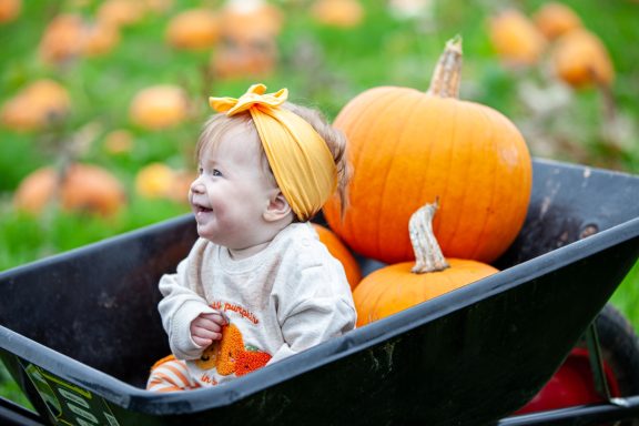 Child in a wheelbarrow with pumpkins, wearing a bright orange headband and smiling.