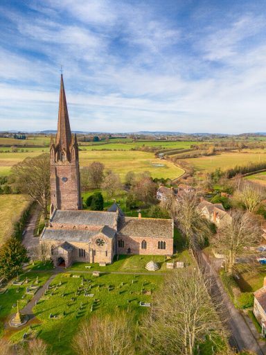 View of a church with a tall steeple surrounded by green fields and trees.