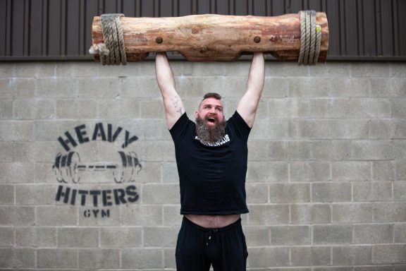 Man in a black shirt hoisting a heavy log overhead, smiling enthusiastically.