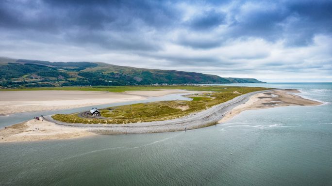Aerial view of a sandy peninsula with a gravel path surrounded by water and green hills.