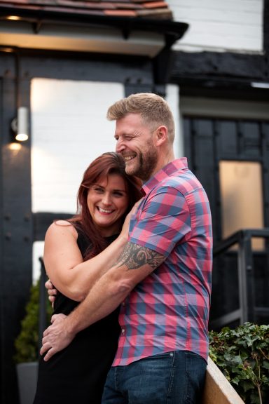 A smiling couple embracing outdoors in casual attire, with a house in the background.