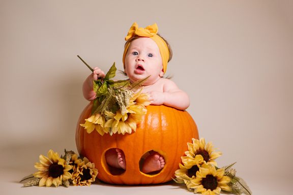 Baby sitting in a pumpkin surrounded by sunflowers, wearing a yellow headband.