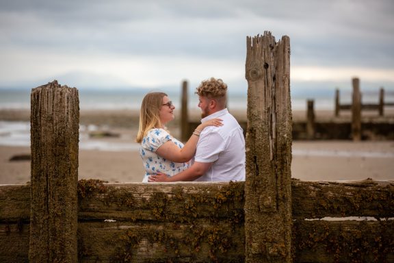 A couple embraces by the beach, framed by weathered wooden posts.