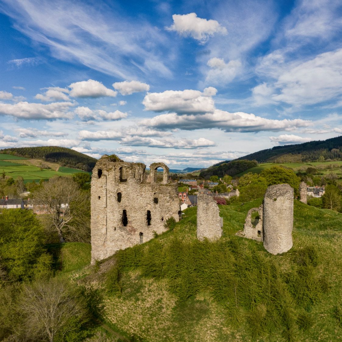 Ruined castle on a hill in Shropshire surrounded by greenery and hills under a cloudy sky.