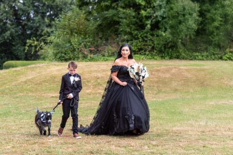 A bride in a black gown and a boy in a suit walking a goat on a lead outdoors.