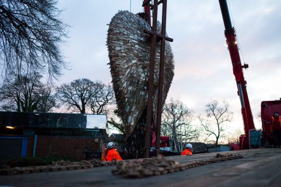Two workers oversee the installation of a large heart-shaped sculpture.