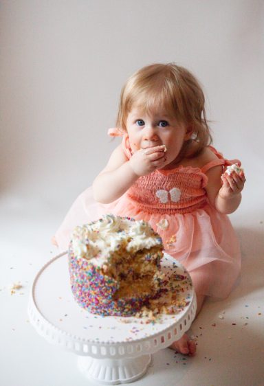 Toddler in a pink dress eating cake, with crumbs around, sitting beside a cake on a stand.