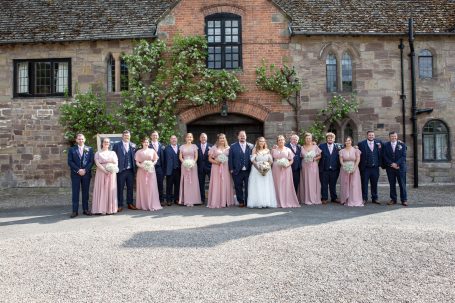 A wedding party poses together in front of a rustic building, dressed in formal attire.