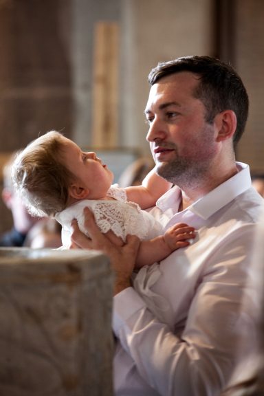 A man holds a baby in a church, both looking at each other fondly.